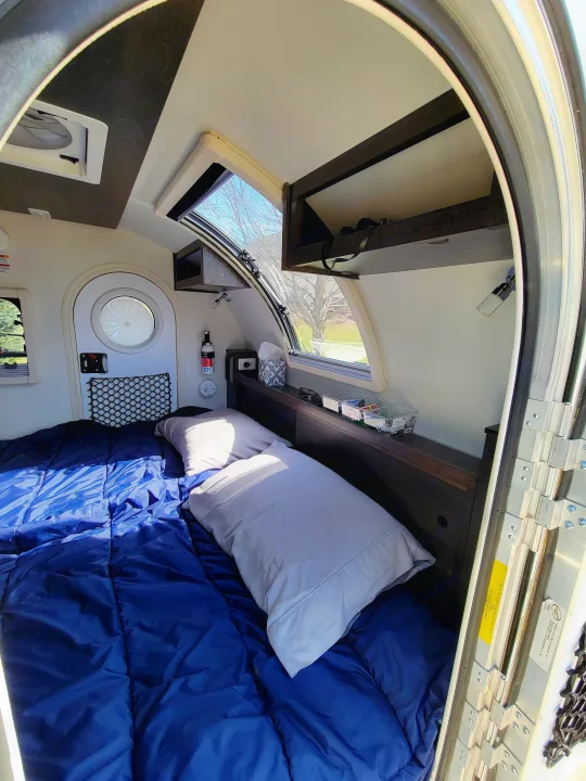 Cozy rv rental camper interior, featuring a bed with a blue comforter and light pillows, overhead storage, and large windows.. nuCamp T@G 2020
