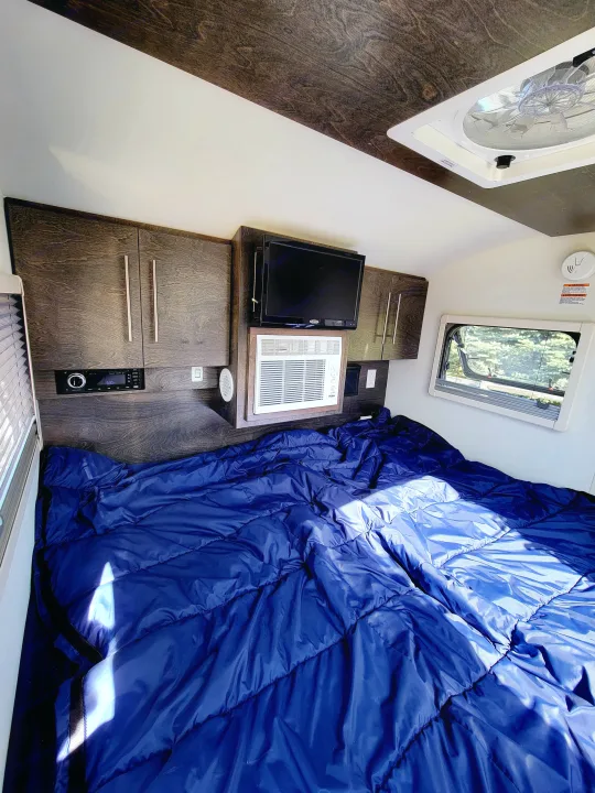 Cozy RV rental bedroom with blue comforter, dark wood cabinets, TV, AC unit, and window view. Perfect for travel.. nuCamp T@G 2020