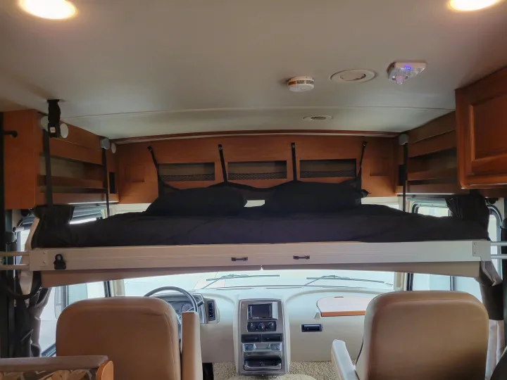 View inside an RV rental: a cozy overhead bunk bed with black bedding sits above the driver's cockpit and dashboard.. Winnebago Vista 2018