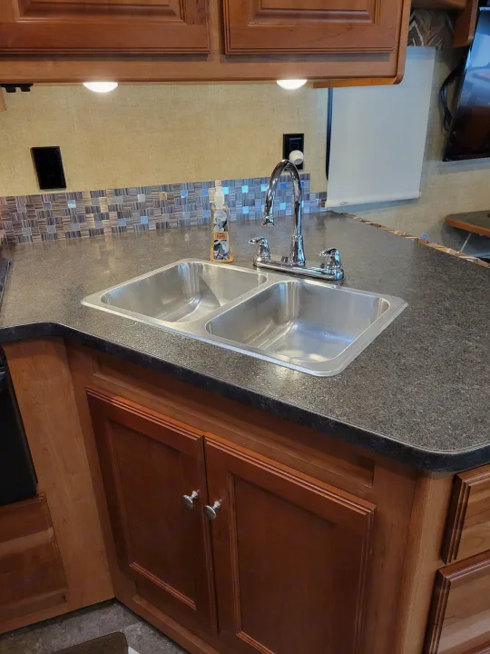 Modern RV rental kitchen with dual stainless steel sink, speckled countertop, tiled backsplash, and warm wood cabinetry.. Winnebago Vista 2018