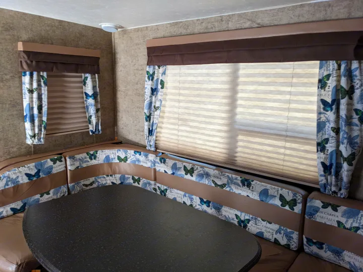 RV rental dinette featuring brown and butterfly-patterned seating, a dark table, and windows with blinds and matching curtains.. Coachmen Freedom Express 2016