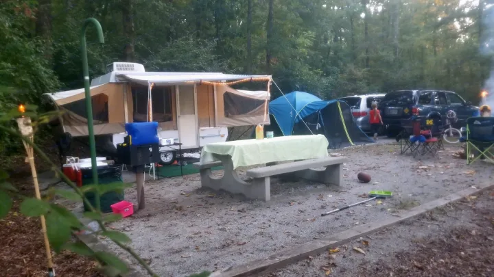 Pop-up camper, like an RV rental, set up with a tent and picnic area at a busy wooded campsite.. Coleman Destiny Royale 1992