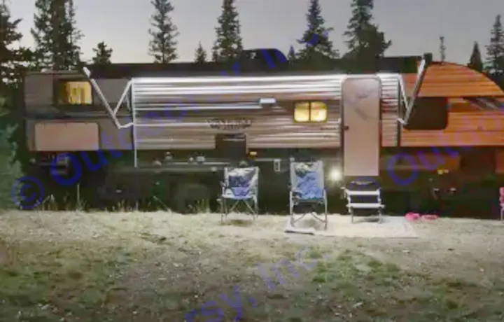 Illuminated RV rental travel trailer at a night campsite. Bright awning lights shine over chairs & a mat, offering a cozy outdoor setting.. Forest River Salem Cruise Lite 2021