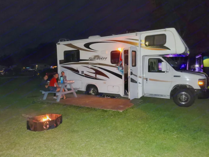 RV rental campsite at night with people gathered around a picnic table by a warm campfire.. Forest River Sunseeker 2013