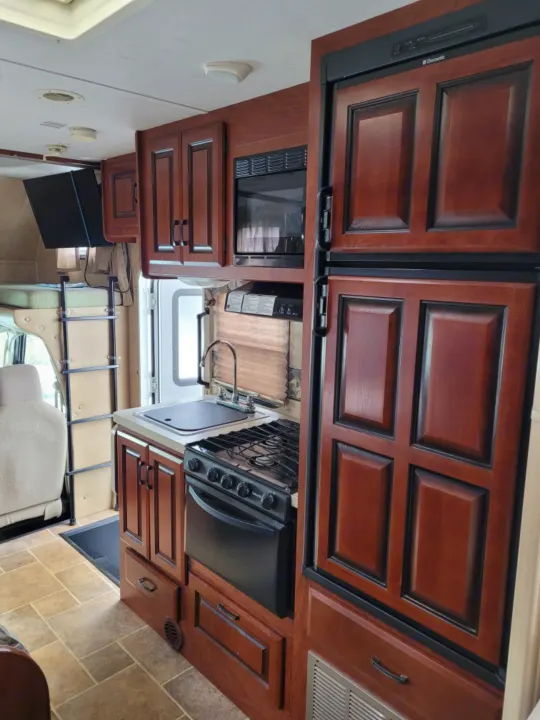 Spacious RV rental interior featuring a full kitchen with microwave, stove, sink, and large fridge, plus a bed.. Forest River Sunseeker 2013