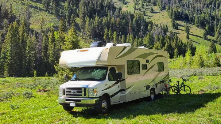 An rv rental parked in a sunny, lush green meadow surrounded by pine trees, with two bikes beside it.. Coachmen Leprechaun 2018