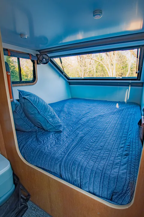 An inviting blue bed inside a compact RV rental, featuring windows with natural views and a ceiling fan.. Campinawe Sport 2022