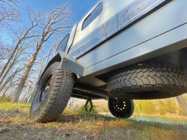 Low-angle shot of a rugged RV rental trailer with large tires and a spare, parked on grass by a serene lake.. Campinawe Sport 2022