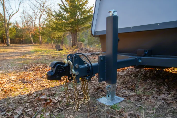 RV rental: The hitch and jack stand of a small camper trailer, parked in a wooded area covered with fallen leaves.. Campinawe Sport 2022