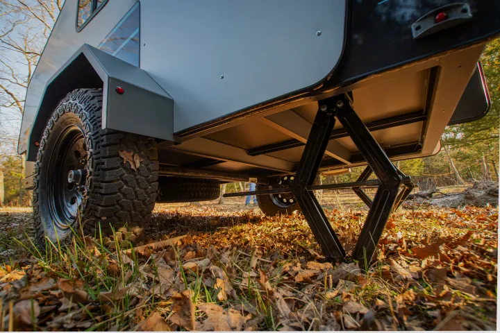 Low-angle view of an off-road travel trailer for RV rental, showcasing its rugged tire and stabilizing jack on autumn leaves.. Campinawe Sport 2022