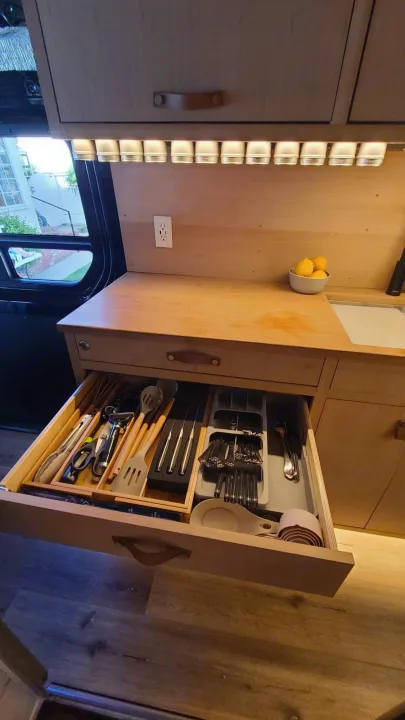 Organized RV kitchen features an open drawer with utensils and a spice rack under bright lights, ideal for your RV rental.. Mercedes-Benz Sprinter 2019