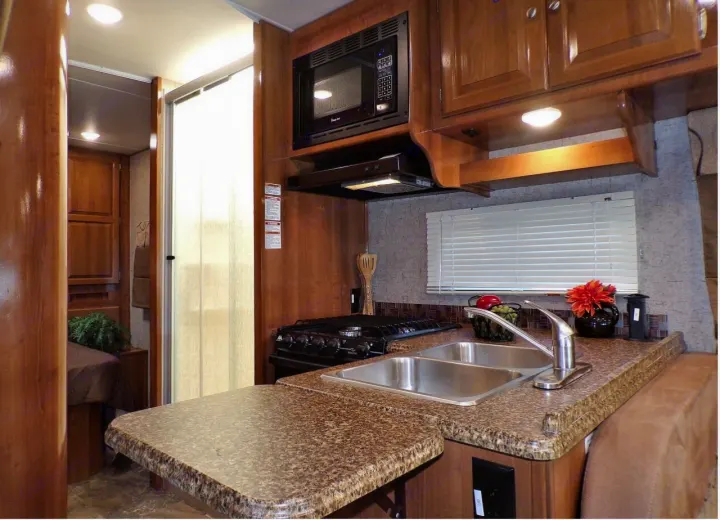 Spacious RV rental kitchen with a double sink, stovetop, microwave, and extendable counter, opening to another room.. Coachmen Leprechaun 2018