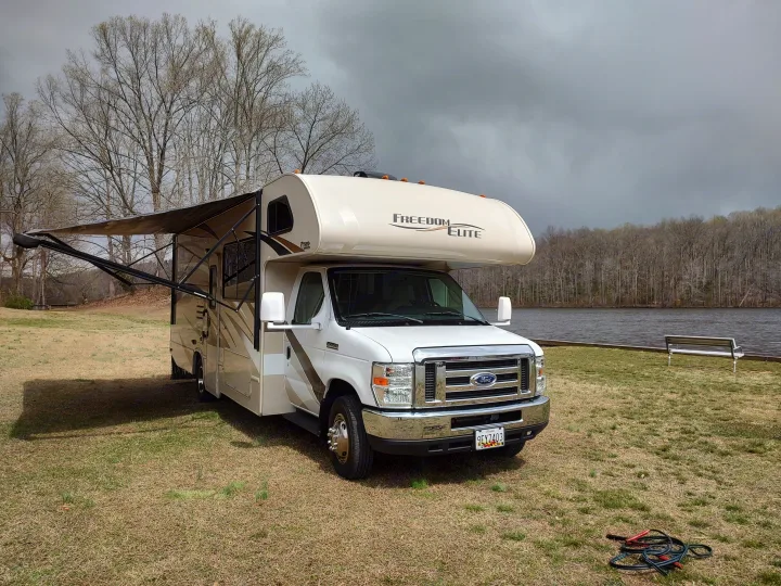 Scenic rv rental parked by a tranquil lake with an extended awning. Perfect for an outdoor adventure under cloudy skies.. Thor Freedom Elite 2017