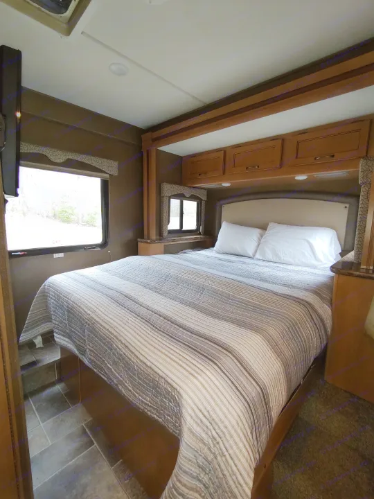 RV rental bedroom interior: a comfortable bed with striped comforter, built-in storage, and an exit door, offering a cozy retreat.. Thor Freedom Elite 2017