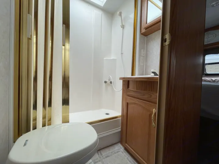 Inside a cozy RV rental bathroom: a white shower stall with gold accents, toilet, and wooden vanity.. Komfort Komfort 2003