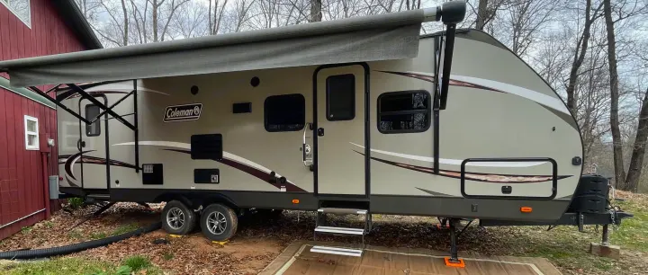 A modern Coleman RV travel trailer, perfect for an RV rental, is parked with its awning extended beside a rustic red building.. Dutchmen Coleman Light LX 2018