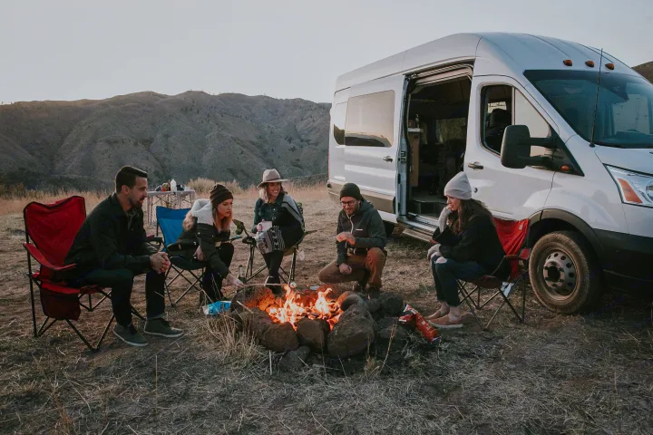 Friends roasting marshmallows by a campfire, with their white RV rental parked in a hilly landscape.. Ford transit 2017