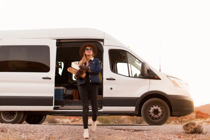 A woman exits a white **RV rental** van with firewood, dressed for camping in a vast desert landscape.. Ford transit 2017