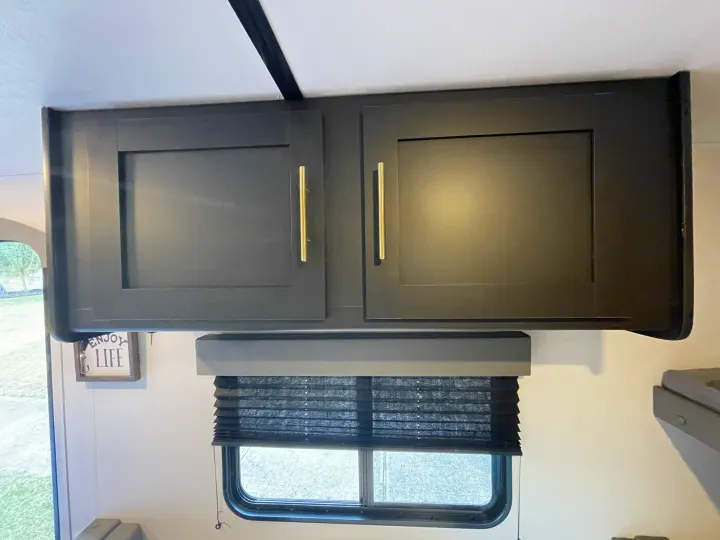 Rv rental interior: dark overhead cabinets, gold handles, and a window with a dark pleated blind. Modern and functional.. Keystone Springdale 2022