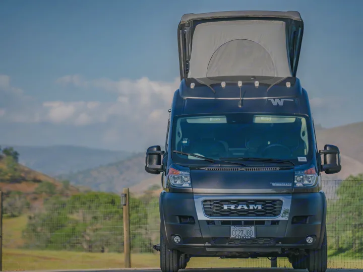 A dark Winnebago Ram ProMaster rv rental van, with its pop-top extended, is parked against a scenic backdrop of hills.. Winnebago Solis 59PX 2023