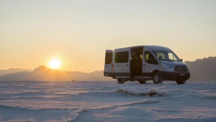 A silver RV rental van with open doors on a textured salt flat at sunset, mountains in background. Perfect for desert travel.. Ford Transit 2017