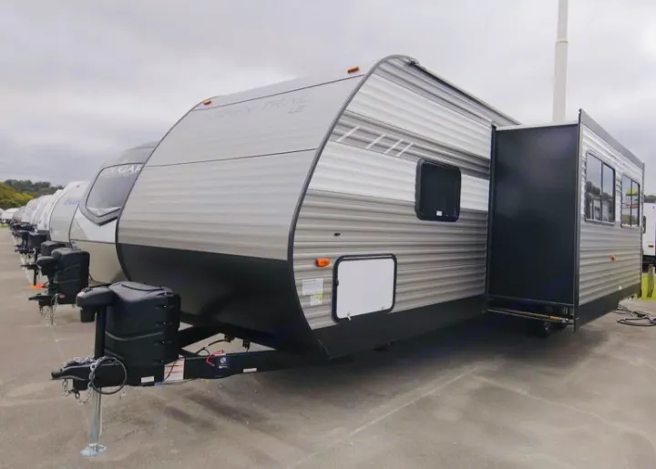 A line of modern travel trailers, including one with an extended slide-out, parked on a lot for rv rental.. Dutchmen Aspen Trail LE 2023
