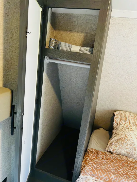 Inside an rv rental: a compact closet with hanging rod and shelf holding neatly folded items, next to the bed.. Dutchmen Aspen Trail LE 2023