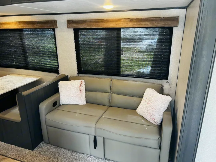 Inside an RV rental: a comfy sofa bed with white pillows and adjacent dinette seating, illuminated by windows with blinds.. Dutchmen Aspen Trail LE 2023