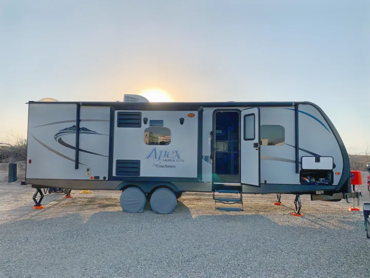 Your next RV rental awaits! A Coachmen Apex travel trailer is parked on gravel, door open, stabilized under a setting sun.. Coachmen Apex 2016