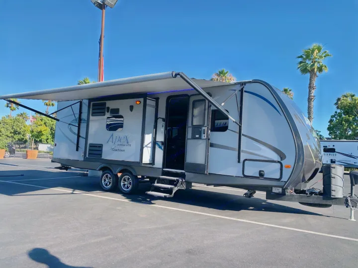 Grey Apex travel trailer with extended awning on an RV rental lot, ready for adventure under a sunny sky.. Coachmen Apex 2016