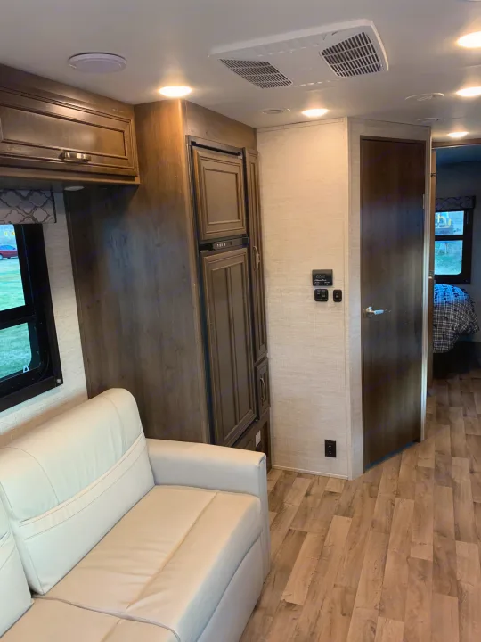 Cozy RV rental interior: view of white sofa, wood-look floor, modern cabinets, AC, and a bedroom beyond.. Jayco Redhawk 2021