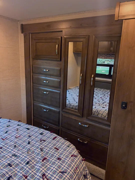 Spacious RV rental bedroom featuring a wooden mirrored wardrobe, dresser drawers, and a cozy plaid bed for comfort and storage.. Jayco Redhawk 2021