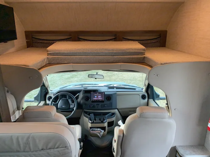 Interior view of an RV rental, highlighting the front cockpit with steering wheel and the cozy bunk bed over the cab.. Jayco Redhawk 2021