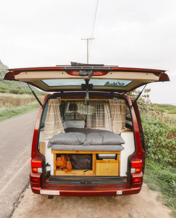 Open tailgate of an RV rental camper van showing its cozy bed and interior storage, parked by a road.. Volkswagen Eurovan 2003