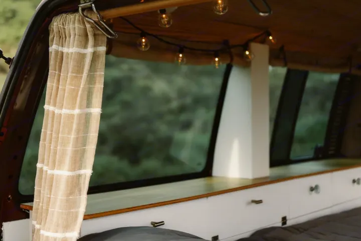 Inside a cozy RV rental: warm string lights, a light curtain, wood details, and a large window overlooking green nature.. Volkswagen Eurovan 2003