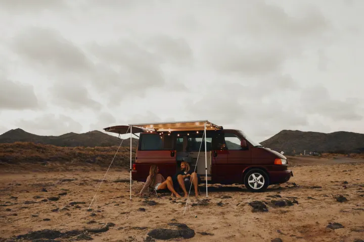 Relaxing rv rental adventure: A couple unwinds in front of their maroon camper van with lights, parked on sandy terrain near mountains.. Volkswagen Eurovan 2003