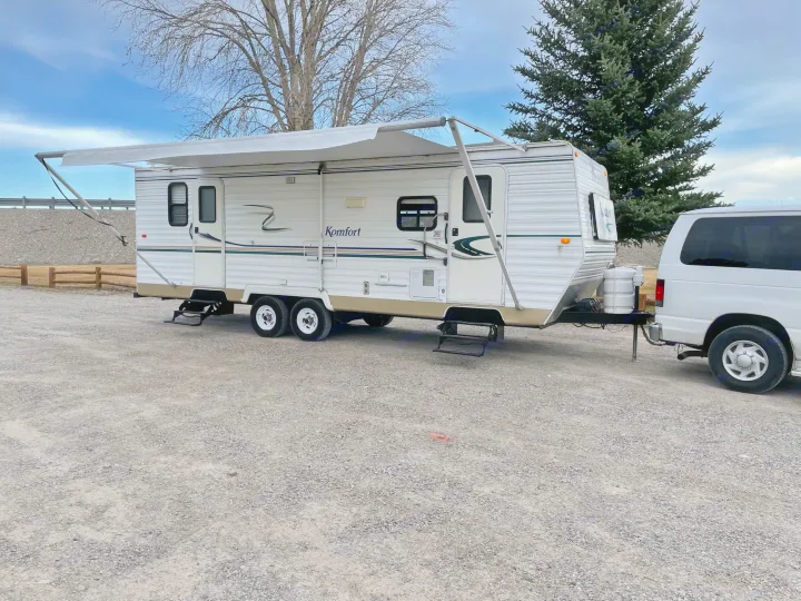 White Komfort travel trailer RV rental with awning extended, hitched to a van on a gravel lot.. Komfort Komfort 2003