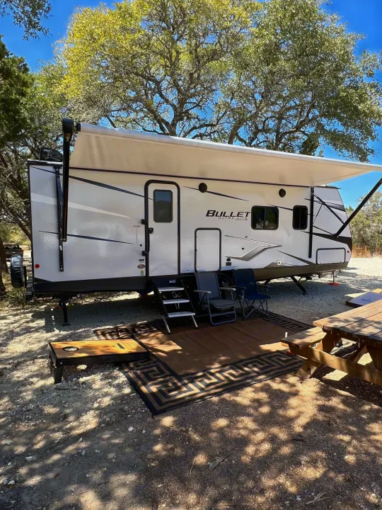 This RV rental is set up for a comfortable outdoor stay, featuring an extended awning, rug, chairs, and a picnic table in a shaded spot.. Keystone Bullet 250BHS 2022