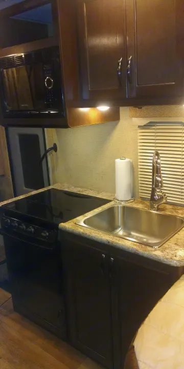 Compact RV rental kitchen featuring dark wood cabinets, speckled countertops, a microwave, stove, and sink.. Thor Freedom Elite 2018