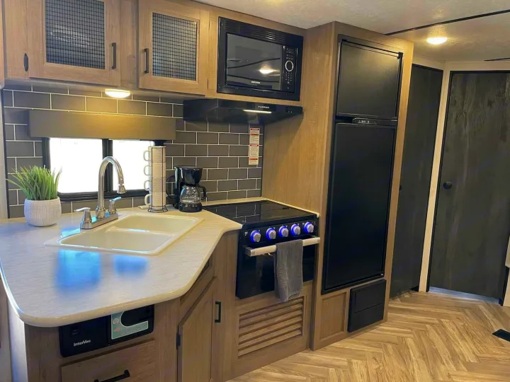Modern RV rental kitchen setup: sleek fridge, microwave, stovetop, and double sink. Light wood cabinets & grey subway tile.. Keystone RV Hideout 2021