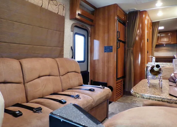 RV rental interior shows a comfortable tan sofa with seatbelts, a dining area, and a kitchen unit with a fridge.. Coachmen Leprechaun 2018