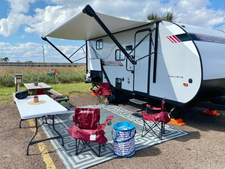 An RV rental campsite setup: white travel trailer with extended awning, outdoor chairs, table, mat, and scenic water view perfect for camping.. Forest River Wildwood 2021