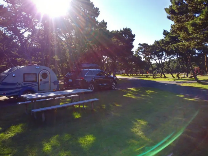 This sunny campground features a teardrop rv rental with an SUV, a wooden picnic table, and tall trees, perfect for camping.. T@G T@G Trailer 2016