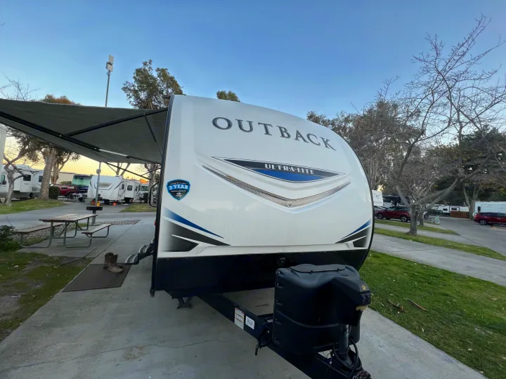 This white Outback travel trailer, possibly an RV rental, is set up at a peaceful campground with its awning extended. Perfect for outdoor adventures.. Keystone Outback 2019