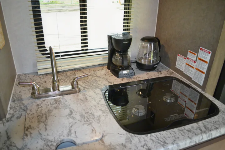 View inside an RV rental showing a compact kitchen setup with a sink, coffee maker, kettle, and covered gas stovetop.. R-Pod R177 2018