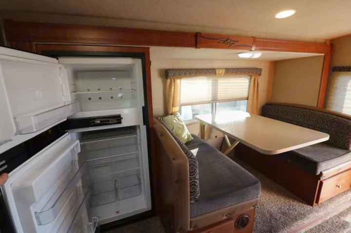 This RV rental shows an open, empty refrigerator beside a dinette with bench seating and a light-colored table.. Adventurer F350 2006
