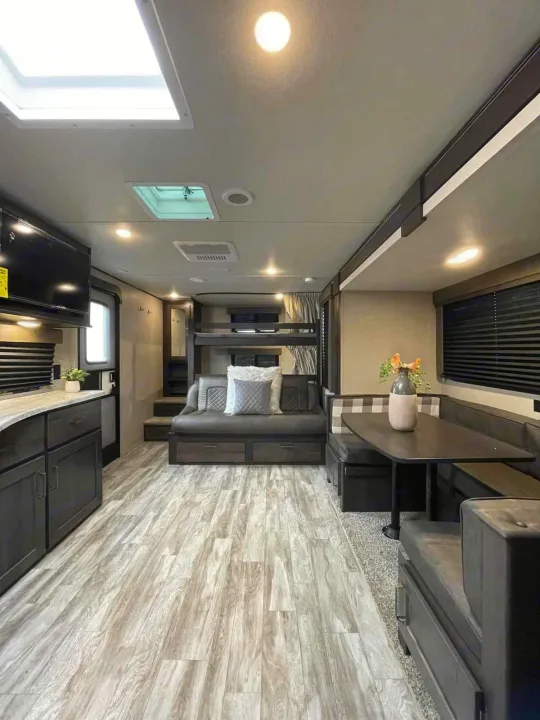 Modern RV rental interior showing light wood floors, dark cabinets, a dining booth, bunk beds, and lounge seating.. Grand Design Transcend Xplor 265BH 2022