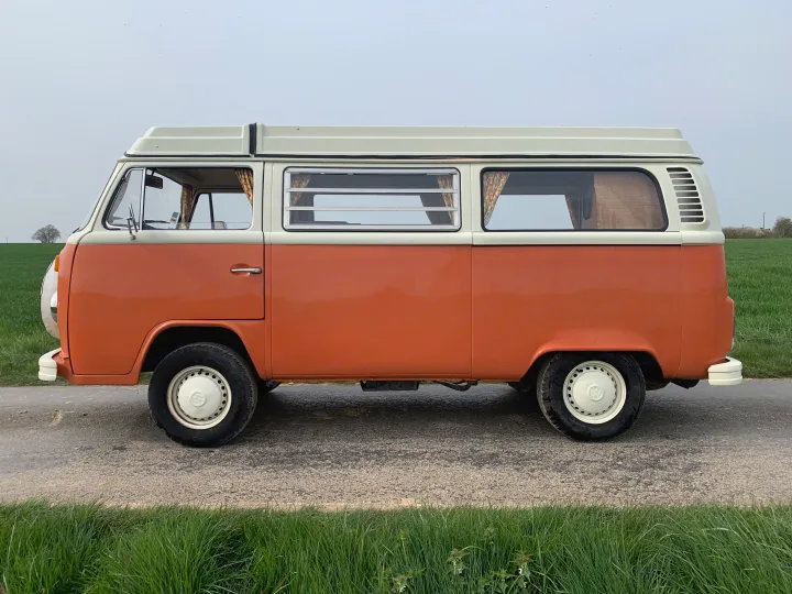 Side profile of a classic orange and white Volkswagen T2 camper van with a high roof, parked on a road. Great for an RV rental.. Volkswagen westfalia 1974