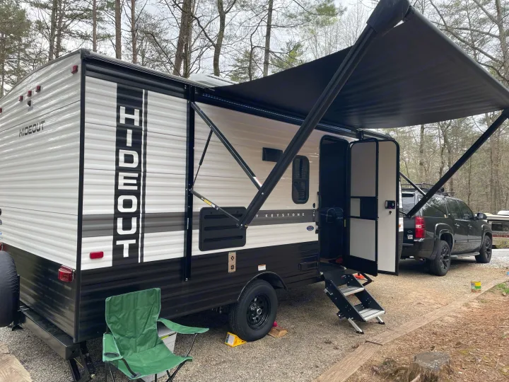 This RV rental is perfectly set up at a wooded campsite with its awning extended and a chair out, ready for relaxation.. Keystone RV Hideout 2022
