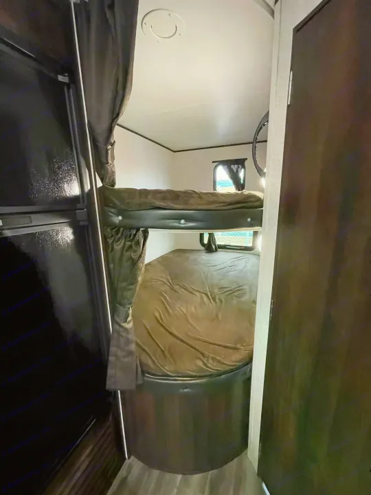 View of an RV rental interior with dark bunk beds & brown bedding, alongside a black refrigerator and dark wooden door.. Jayco Rocky Mountain 242BHSW 2019